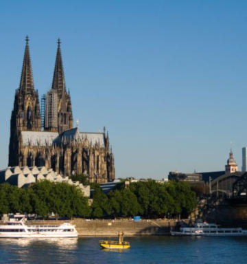Der Kölner Dom unter blauem Himmel, vorn der Rhein mit Schiffen, rechts die Hohenzollernbrücke